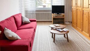 Un soggiorno con un divano rosso, un tavolino rotondo con riviste, un televisore su un mobile in legno e un'ampia finestra con tende.