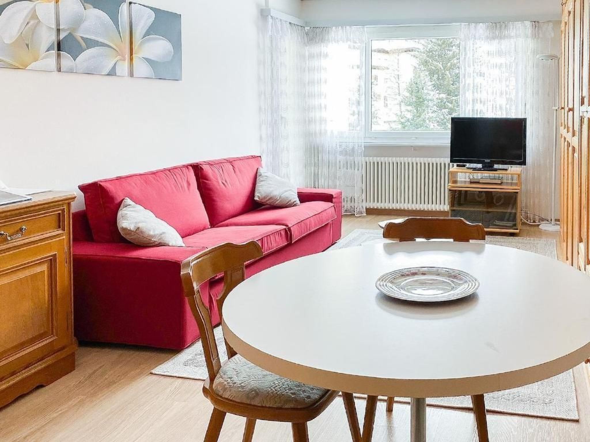 En stue med en rød sofa, et træskab, et hvidt bord og et fjernsyn på et træstel.