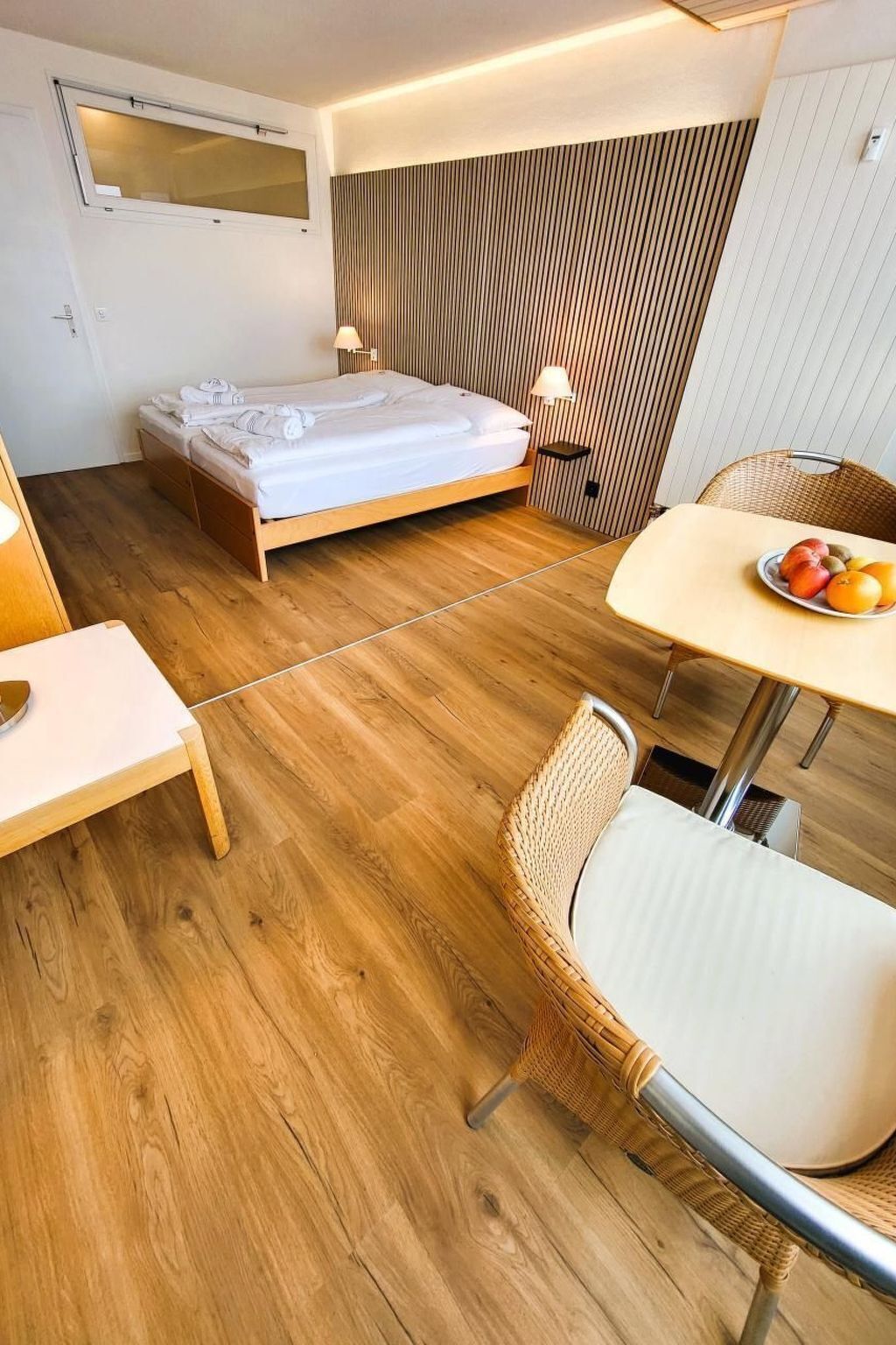 A modern hotel room with a wooden floor, a bed, a small table with fruit, and a wicker chair.