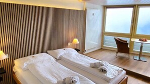 Et moderne hotelværelse med to senge, trægulv, et stort vindue med udsigt og et lille bord med stol.