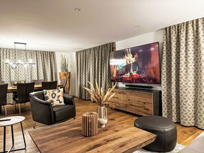 A modern living room with a flat-screen TV, leather sofa, wooden coffee table, and a dining area with a chandelier.