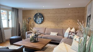A cozy living room with a wooden wall, a round clock, a sofa, a coffee table, and a window with curtains. - Casa Ladina 4 by Interhome (Falera)