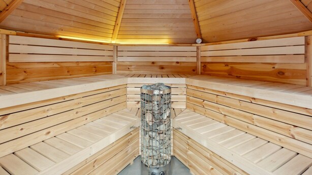 Eine Sauna mit Holzbänken, die in einer Halbkreisform angeordnet sind, einem zentralen Steinofen und einer geneigten Holzdach.