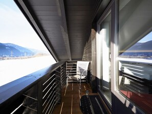 Ein schmaler Balkon mit Metallgeländer, einem Stuhl und einem Fenster mit Blick auf eine verschneite Berglandschaft.