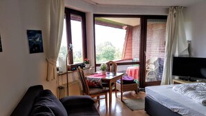 Ein Zimmer mit einer Couch, einem Bett, einem Fernseher, einem Esstisch mit Stühlen und einem Balkon mit Blick auf Bäume.