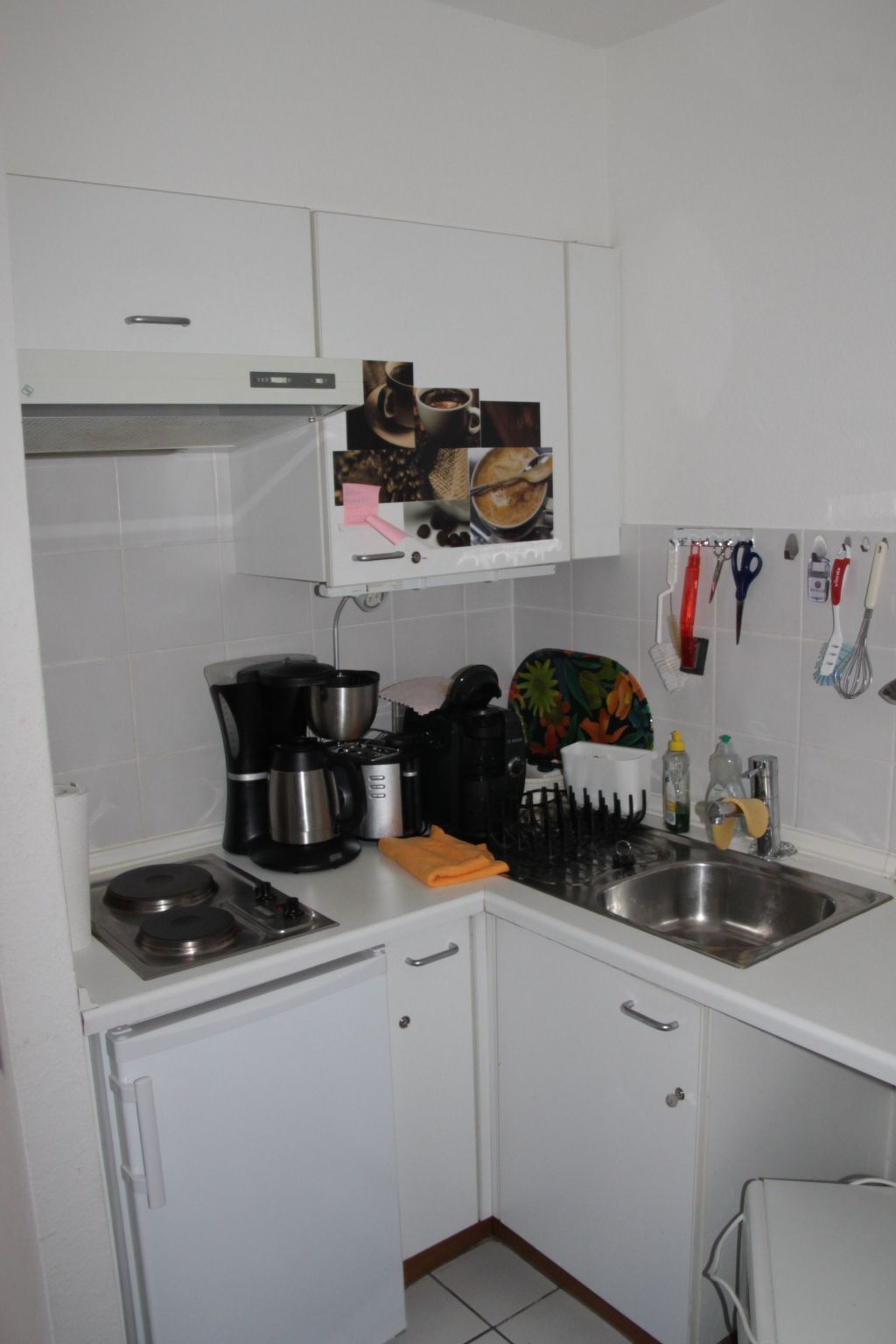 A compact kitchen with white cabinets, a stainless steel sink, and various appliances including a coffee maker and a kettle.
