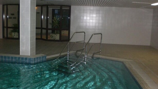 Ein Hallenschwimmbad mit klarem Wasser, Metallleitern, gefliesten Wänden und einer Zimmerpflanze im Hintergrund.