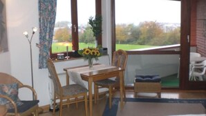 Ein gemütliches Zimmer mit Holztisch, zwei Stühlen, einer kleinen Couch und Blick auf eine grüne Landschaft durch große Fenster.