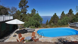 Una piscina con sillas de descanso y una mesa, rodeada de baldosas de piedra y vegetación, con vista al mar y árboles al fondo.