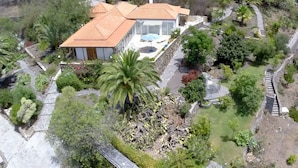 Vista aérea de una casa con techo de tejas, rodeada por un jardín ajardinado con palmeras y arbustos variados.