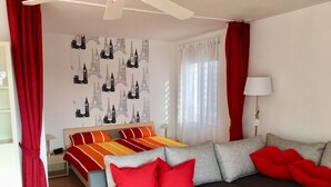 Une chambre d’hôtel moderne avec un lit, un canapé gris, une literie à rayures rouges et oranges, et un mur recouvert d’un papier peint représentant un paysage urbain.