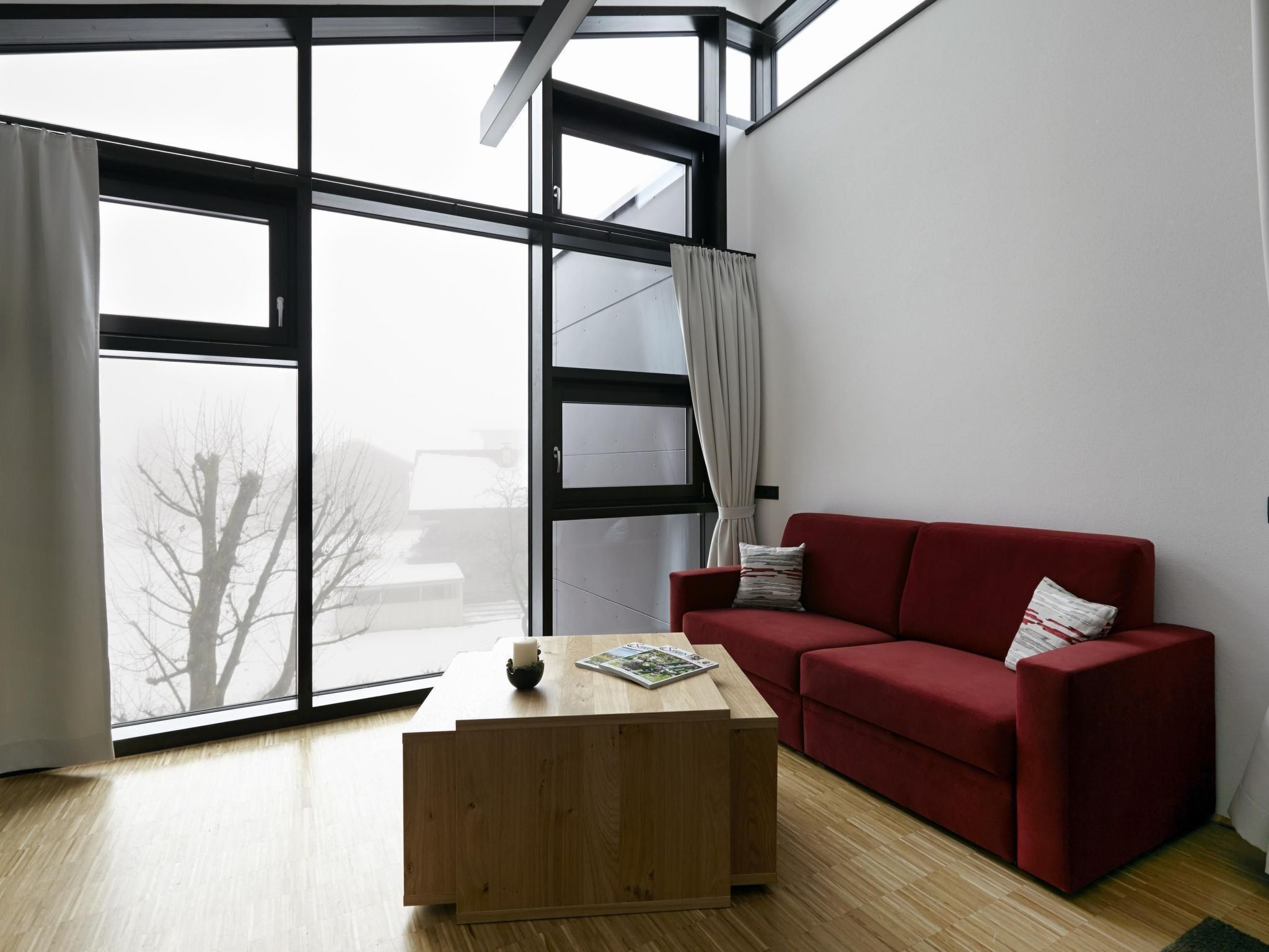A modern living room with a red sofa, a wooden coffee table, and large windows offering a view of the outside.