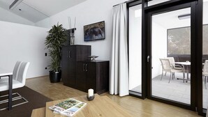 En moderne stue med trægulv, et spisebord med stole, et mørkt træskab, en potteplante, en hvid gardin og en glasdør, der fører til et udendørs område.