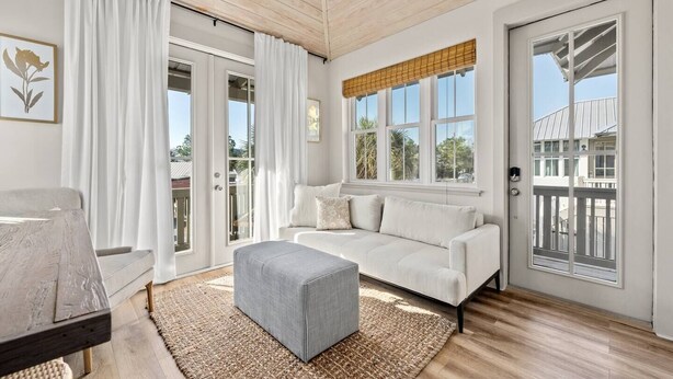 A modern living room with a white sofa, a grey ottoman, and a large window with a view of the outside.