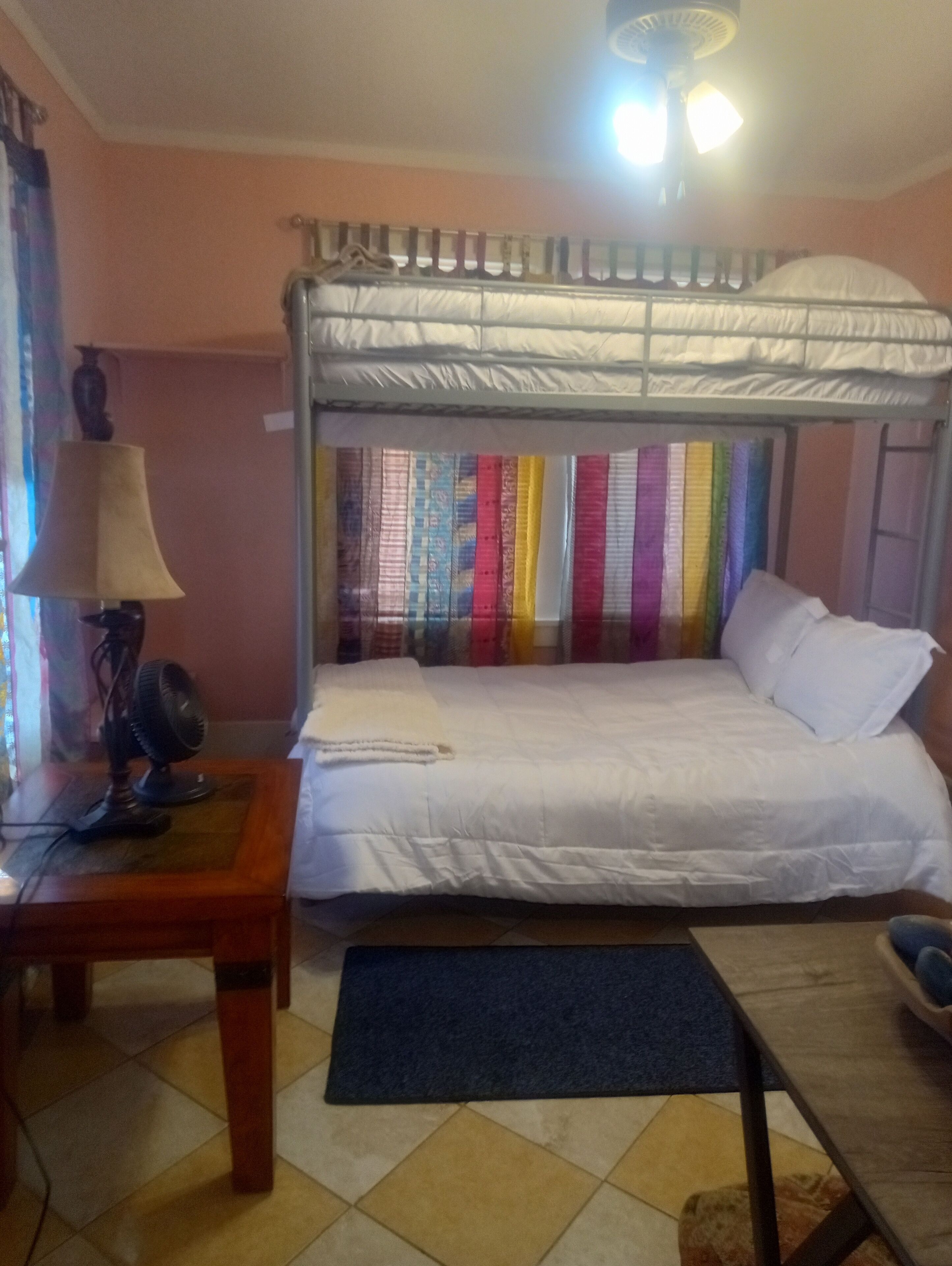 Um quarto com uma cama de solteiro e beliche, uma escrivaninha com luminária e um ventilador.