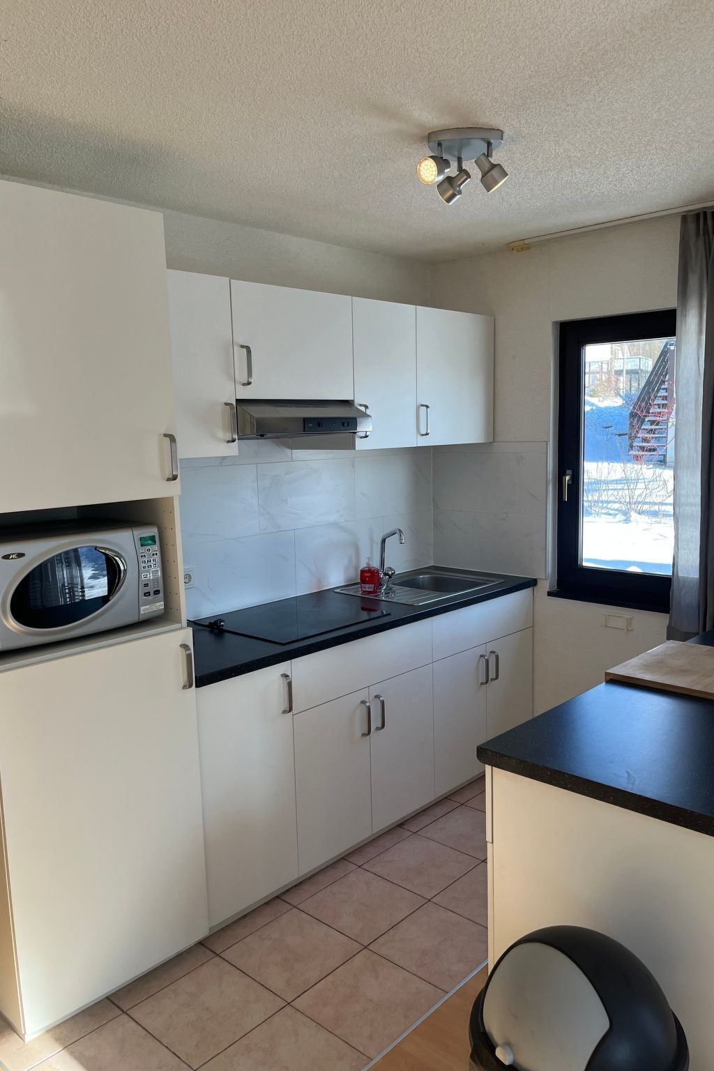 Una cocina compacta con armarios blancos, una encimera negra, microondas, un lavabo y una ventana con vistas a un edificio.