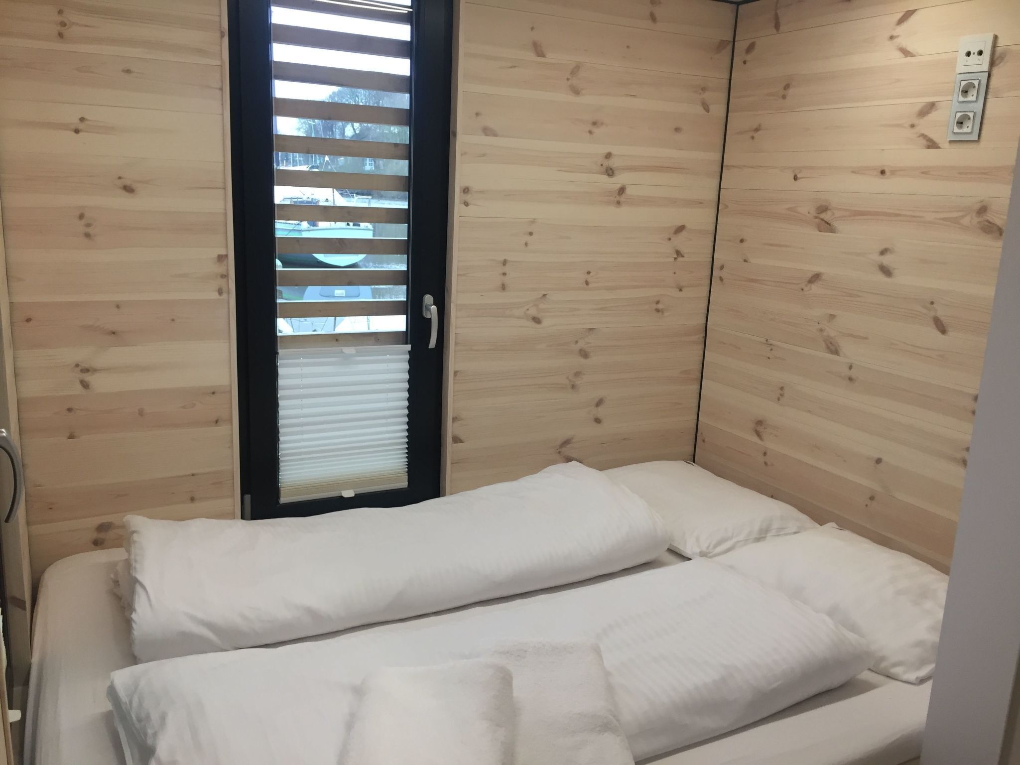 Una habitación pequeña y acogedora con paredes de madera, una cama individual con ropa blanca y una ventana con persianas.