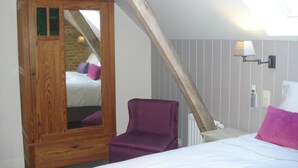 Ein Schlafzimmer mit schrägverlaufender Decke, einem Holzschrank, einem violetten Sessel und einem Bett mit weißer Bettwäsche.