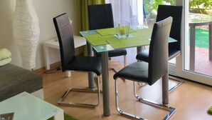 Un comedor con una mesa de vidrio y cuatro sillas, un armario blanco con puertas de cristal, y vista a zonas verdes exteriores.