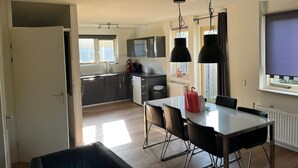 Une cuisine moderne avec des meubles de couleur sombre, un plan de travail blanc et un coin repas comprenant une table en verre et des chaises noires.