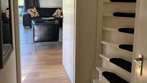 Un intérieur moderne avec un escalier menant à un deuxième étage, un espace de vie comprenant un canapé et une table basse, et une vue sur l’extérieur par une fenêtre.