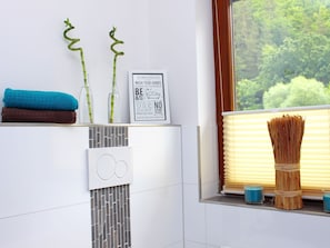Une salle de bain équipée d’un porte-serviettes où sont posées deux serviettes, d’un panneau encadré affichant les règles de la salle de bain, d’une fenêtre avec des stores et d’un panier décoratif contenant des roseaux.