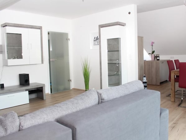 Un salon moderne avec un canapé gris, un meuble de rangement blanc pour les appareils électroniques et une porte en verre donnant sur une salle de bain.