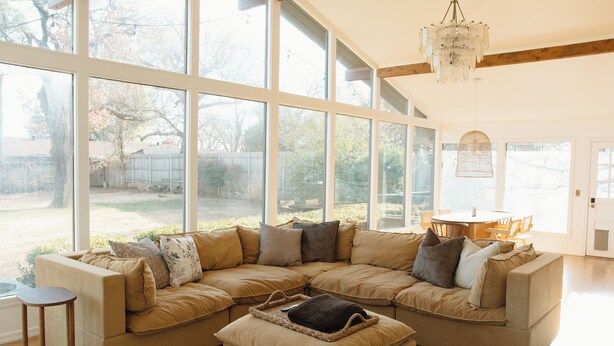 A large L-shaped sectional sofa with multiple cushions, a small side table, and a woven ottoman in a bright room with large windows and a chandelier. - NEW Boutique Historical Home Downtown Edmond (Edmond)
