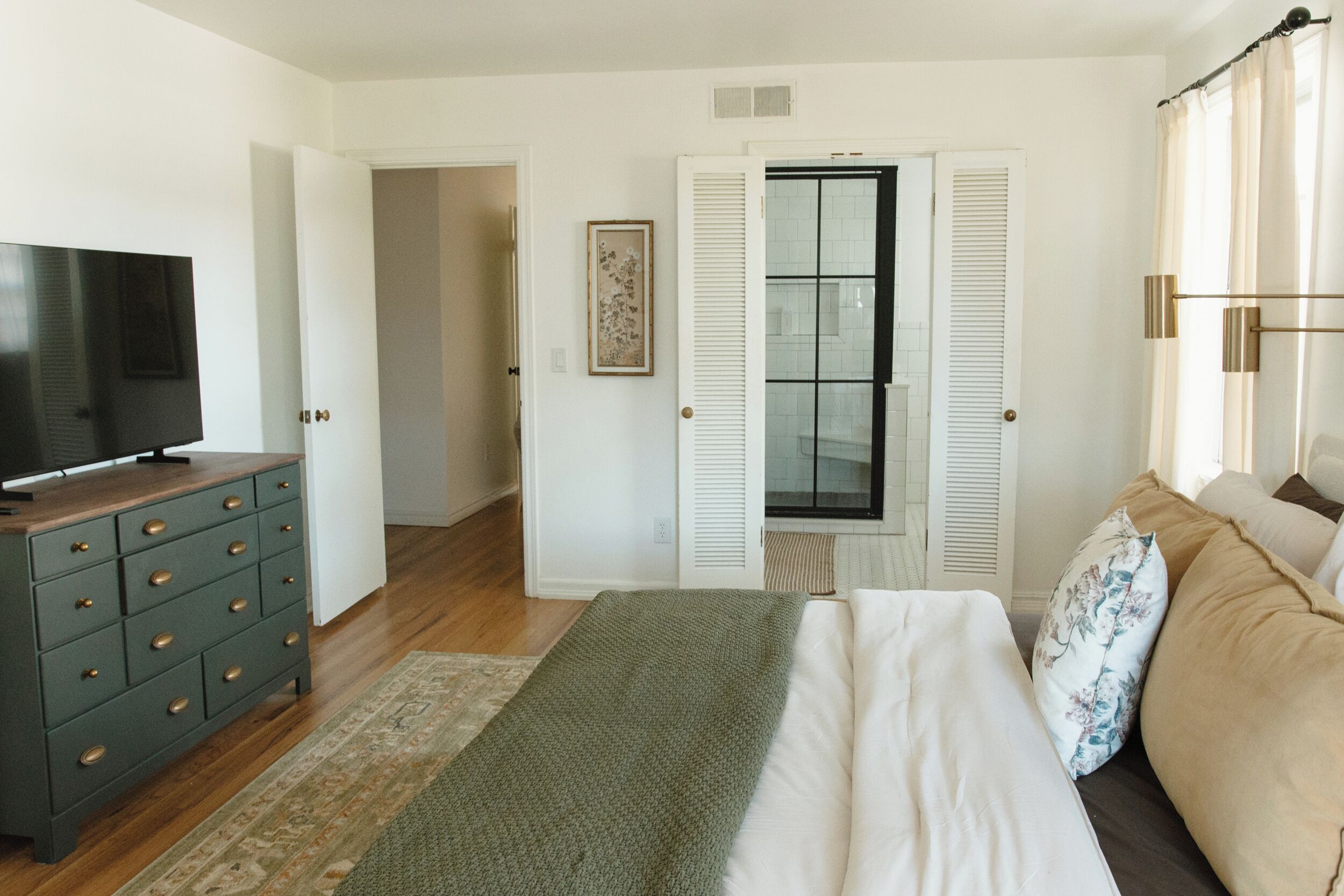 Un dormitorio con cama, cómoda, televisor y ventana con contraventanas.