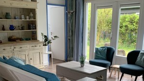 A modern living room with a white sofa, a coffee table, and a blue armchair. There is a wooden cabinet with decorative items and a view of greenery outside. - mit Garten, Grill und Terrasse by Interhome (Julianadorp aan zee)