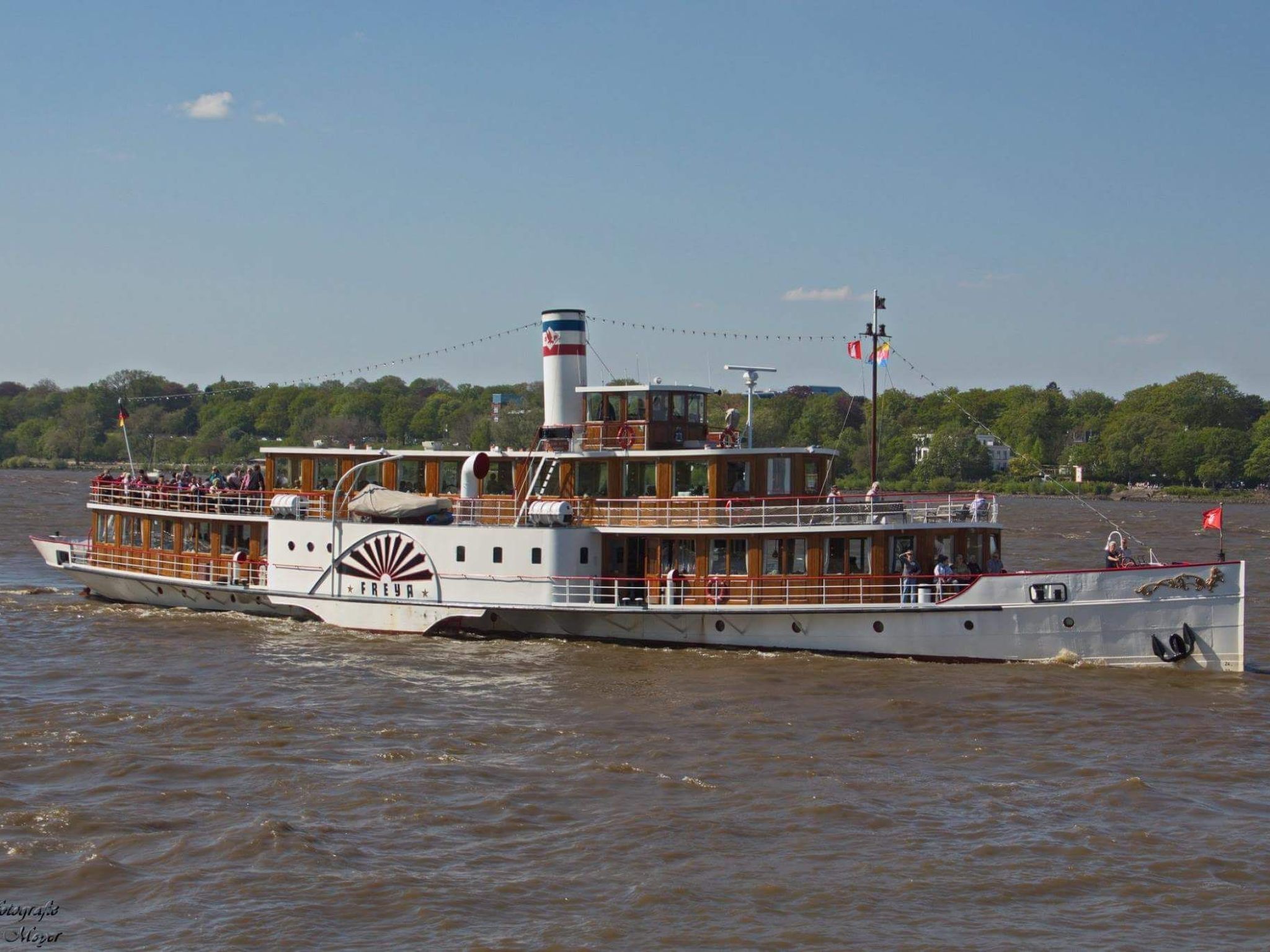 Een rivierboot met meerdere dekken en een opvallende schoorsteen, die op een rivier vaart.