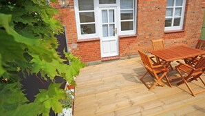 A brick house with two white-framed windows, a wooden deck with a table and chairs, and a vine-covered wall.
