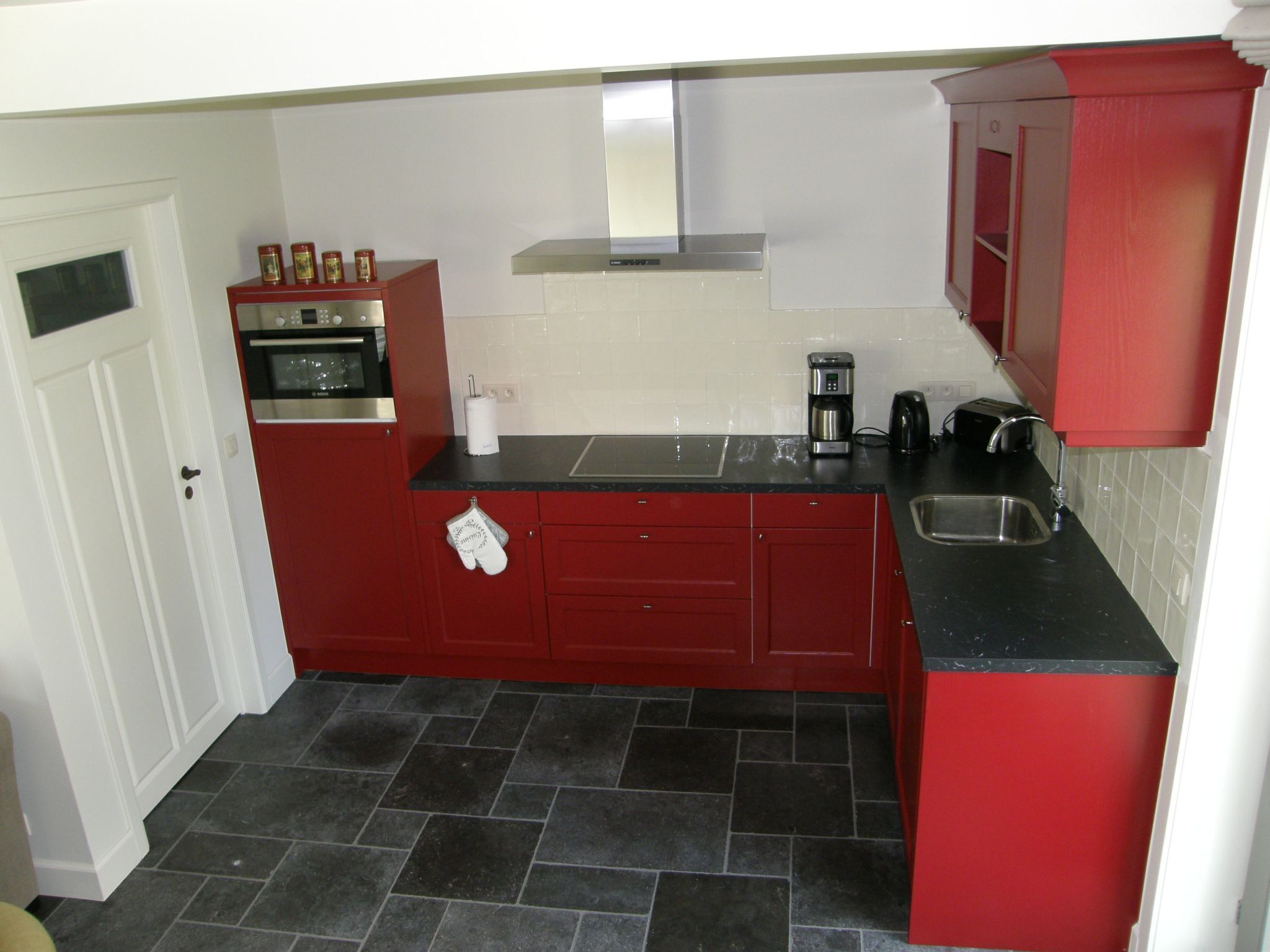 Una cocina compacta con armarios rojos, una encimera negra, un horno de acero inoxidable, una cafetera y un lavabo.