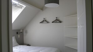 Et soveværelse med skråvæg, en seng med hvidt sengetøj, en hængende lampe og en reol fastgjort til væggen.