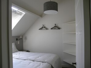 Ein Schlafzimmer mit schrägen Wänden, einem Bett mit weißer Bettwäsche, einer Hängelampe und einem Wandregal.