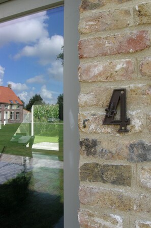 Eine Ziegelwand mit einer Nummer '4' als Schild, ein Fenster das ein Gebäude und Grünflächen reflektiert, und ein klarer Himmel.