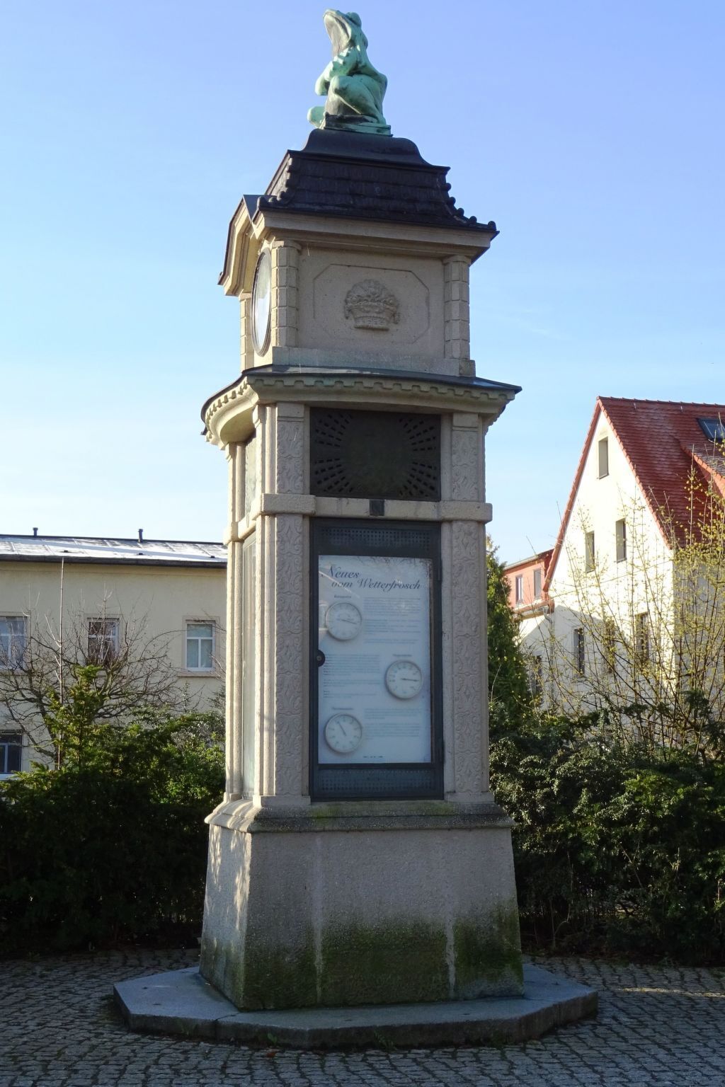 Un monument en pierre orné d’une horloge et d’une plaque commémorative, entouré d’un pavage en pierre et de verdure.