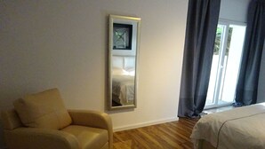Ein Hotelzimmer mit Holzboden, einem beigefarbenen Sessel, einem Bett mit weißer Bettwäsche, einem großen Spiegel an der Wand und einem Fenster mit Vorhängen.