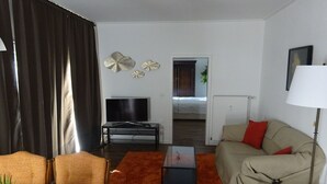 Ein Wohnzimmer mit Fernseher, Sofa, Couchtisch und Blick ins Schlafzimmer.