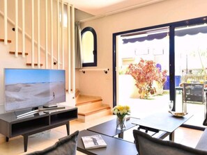 Ein modernes Wohnzimmer mit Flachbildfernseher, Couchtisch und Sofa. Eine Treppe führt zu einem Außenbereich mit Blick auf einen Garten und ein Gebäude.