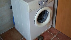 Una lavadora blanca y compacta con panel de control y tambor de carga frontal, ubicada en una esquina con paredes y suelo de baldosas.