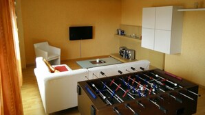 Una sala de estar acogedora con mesa de fútbol, un sofá blanco, televisor de pantalla plana y una zona de cocina con cafetera.