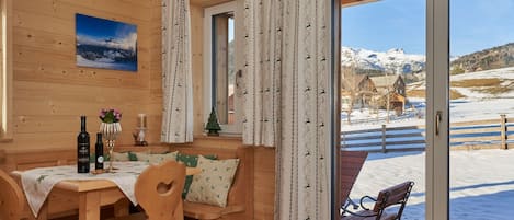 A wooden interior with a dining table, chairs, and a view of a snowy landscape through a glass door.