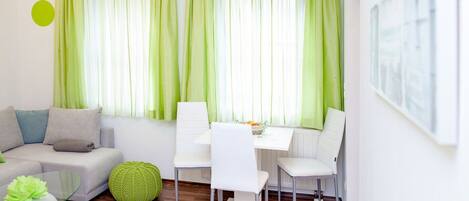 Una sala de estar moderna con un sofá blanco, cortinas verdes y un comedor con sillas blancas y una mesa.