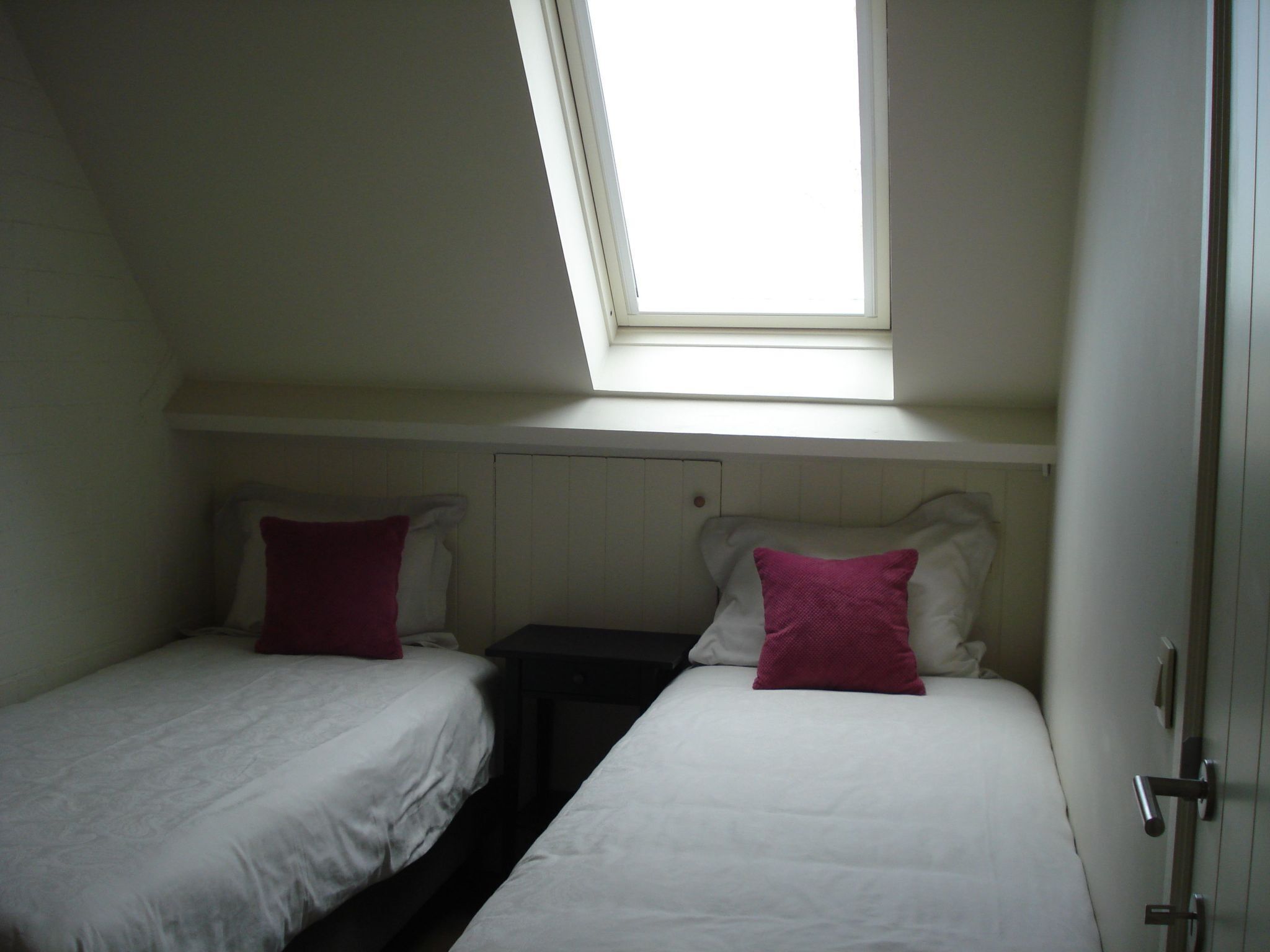 A small, dimly lit room with two single beds, each with a pillow and a nightstand. A skylight provides natural light.