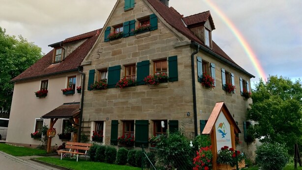 Edifici de pedra amb persianes verdes i jardineres florals, una banca de fusta i un cartell que indica un restaurant.