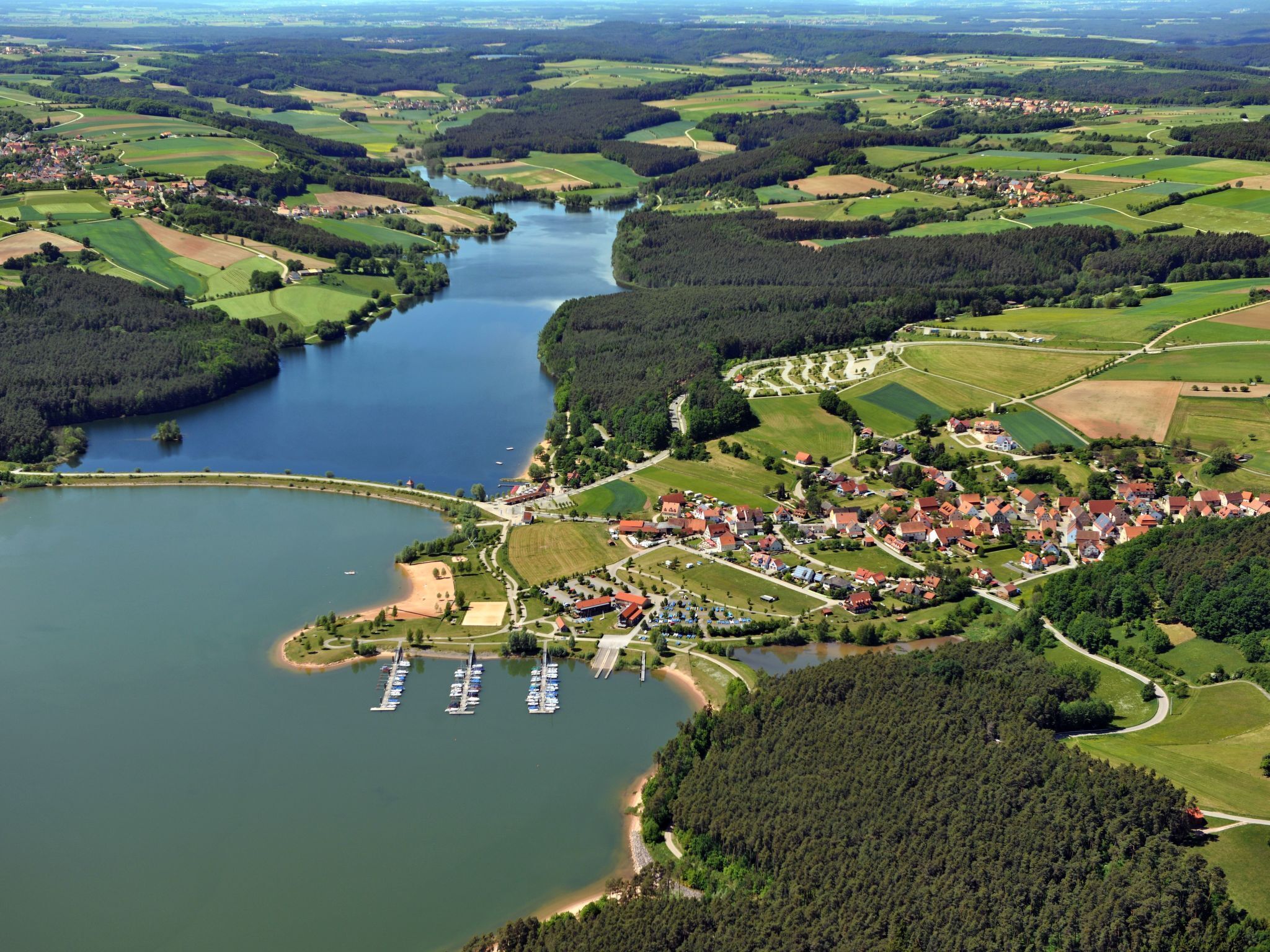 Vista aérea de uma vila à beira de um lago, com um porto e cercada por campos e florestas.