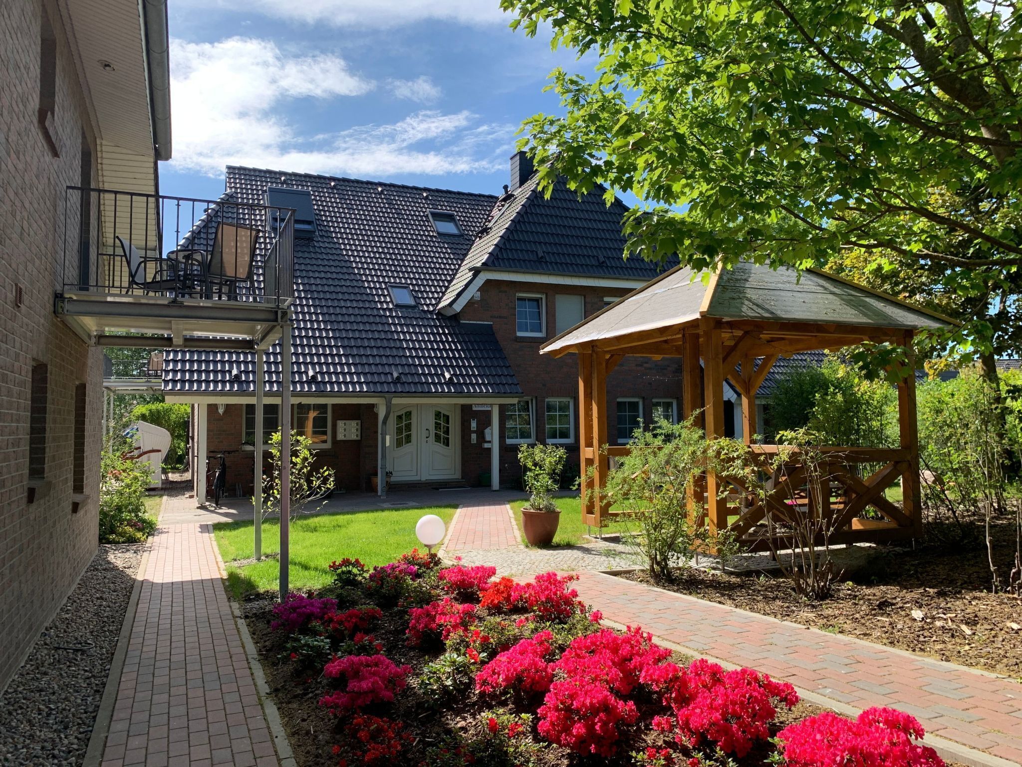 Ein Innenhof mit einem Backsteinhaus, einer Holzlaube und farbenfrohen Blumenbeeten.