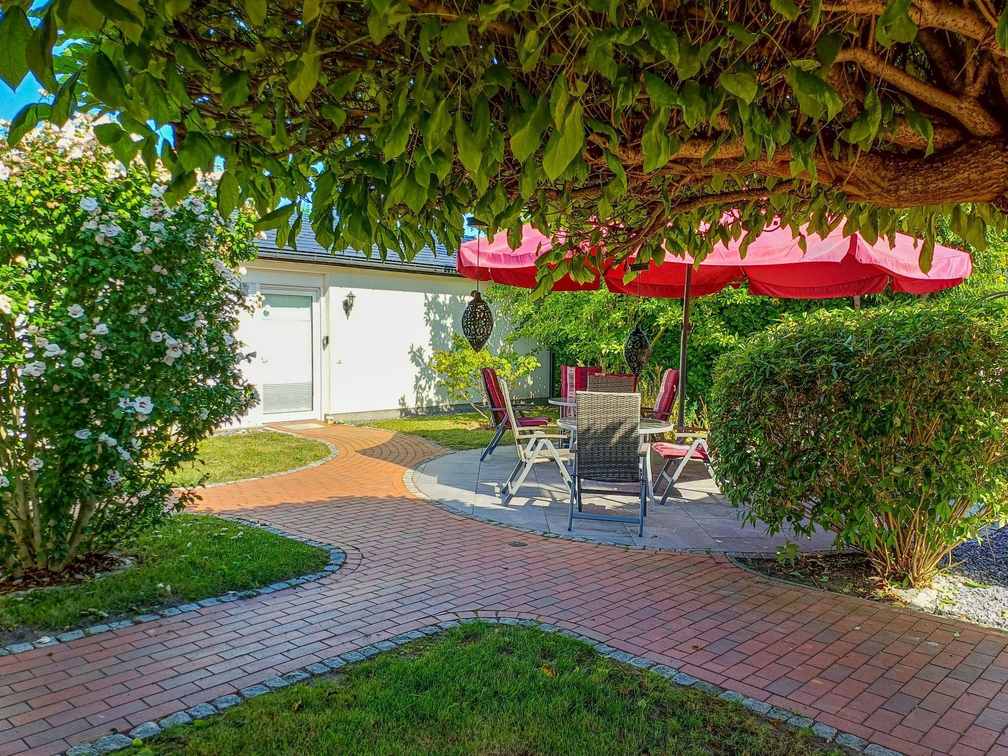 Een patio met een geplaveid pad van stenen, tuinmeubels en een overdekt zitgedeelte met rode parasols.