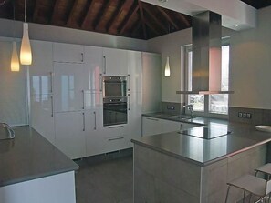 Eine moderne Küche mit weißen Schränken, einem eingebauten Backofen, einer großen Kochinsel mit Spüle, Pendelleuchten und einem Fenster mit Jalousien.